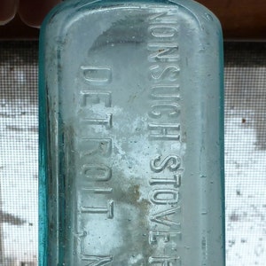 Puede incluir: Una botella de vidrio transparente con un tono azul. La botella tiene una etiqueta que dice "NONSUCH STOVE POLISH DETROIT MICH."