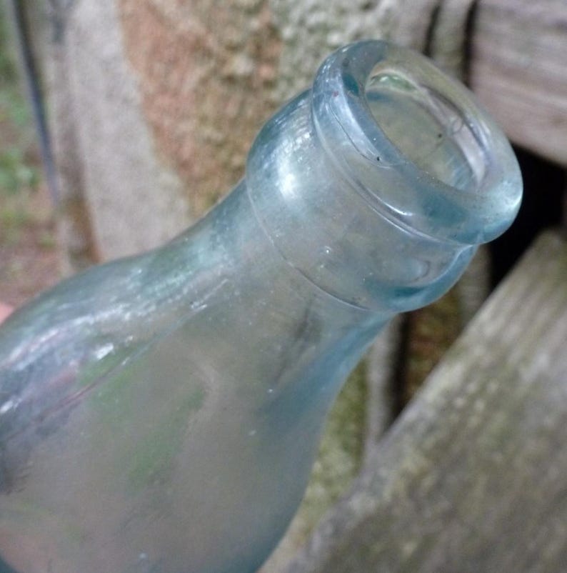 Puede incluir: Una botella de vidrio transparente y antigua con cuello redondeado y labio ensanchado. La botella tiene un ligero tinte azul y parece envejecida. La superficie de la botella muestra signos de desgaste, lo que sugiere que es vintage.