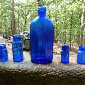 Peut inclure: Cinq bouteilles anciennes en verre bleu cobalt de tailles variées sont disposées en rangée. Les bouteilles sont exposées sur une surface texturée, avec une toile de fond d'arbres et de feuillage.