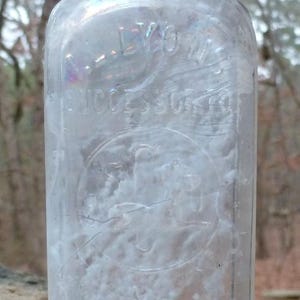 May include: A clear, rectangular glass bottle with a rounded top and a short neck. The bottle has embossed text, including the words "LYON" and "SUCCESSORS". The bottle appears to be an antique.