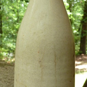 May include: A vintage, off-white ceramic bottle with a rounded body and a slightly flared top. The bottle has a smooth, slightly textured surface and appears to be hand-crafted.