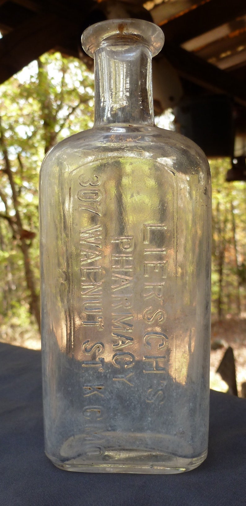 May include: A clear glass bottle with a textured surface. The bottle has the text "LIERSCH'S PHARMACY 307 WALNUT ST. K.C." etched into the glass.