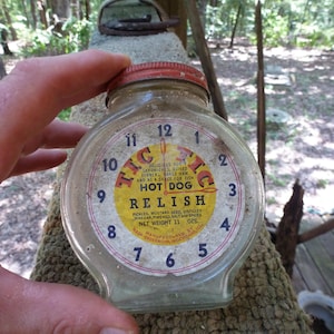 Puede incluir: Un tarro de vidrio vintage con tapa roja, con un diseño de esfera de reloj con las palabras "HOT DOG RELISH". La etiqueta incluye los ingredientes y la información del fabricante. El tarro se sostiene frente a un fondo borroso al aire libre.