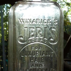 Può includere: Bottiglia di vetro trasparente con testo in rilievo. La bottiglia riporta "WINARICK'S JERIS REC'D" in alto, con "FOR LUXURIANT HAIR AND CLEAN SCALP" in un cerchio centrale. In basso si legge "PREPARED BY AR WINARICK NEW YORK".