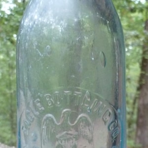 Puede incluir: Una botella de vidrio azul claro vintage con texto en relieve y un emblema de &aacute;guila. La botella tiene forma redondeada y cuello estrecho. El texto dice "Eagle Bottling Co. Registered Kansas City, Mo."