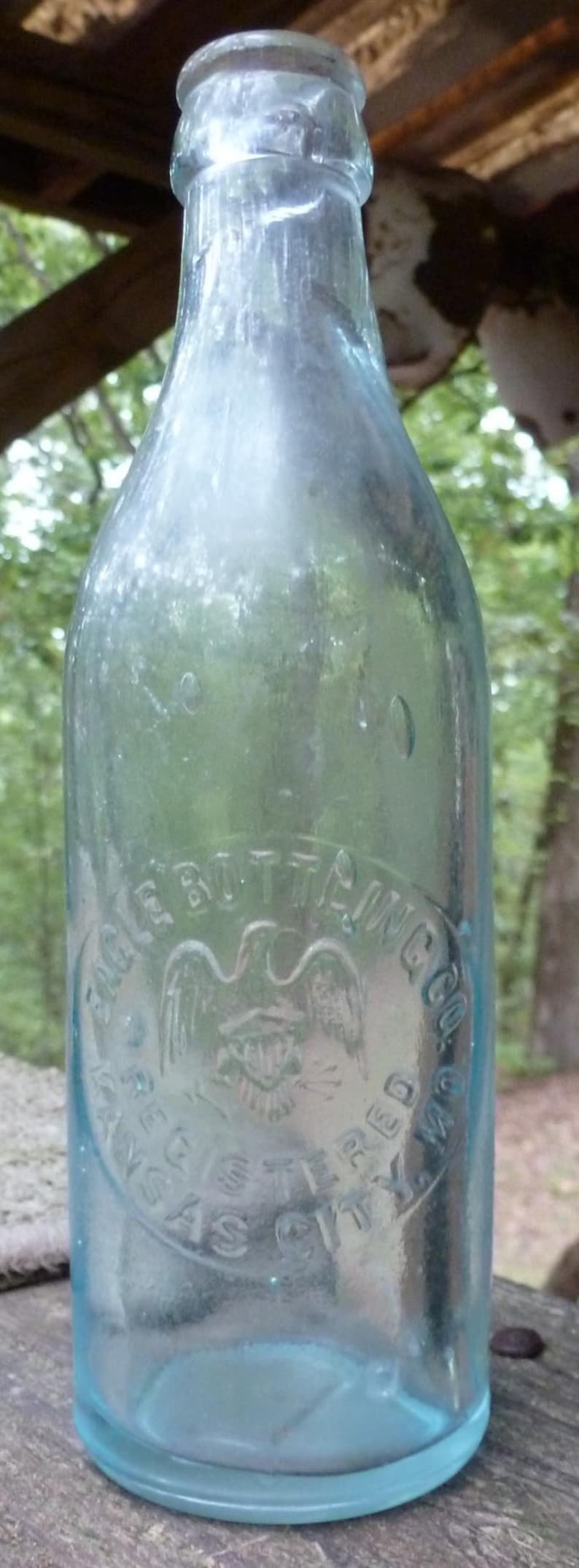 Puede incluir: Una botella de vidrio azul claro vintage con texto en relieve y un emblema de &aacute;guila. La botella tiene forma redondeada y cuello estrecho. El texto dice "Eagle Bottling Co. Registered Kansas City, Mo."
