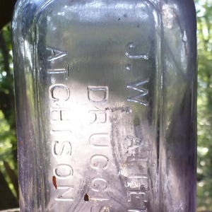 May include: A vintage, rectangular, purple glass bottle with a rounded shoulder and a short neck. The bottle has embossed text that reads "J.W. Allen & Co. Druggists, Atchison, Kans."