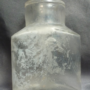 Peut inclure: Une bouteille en verre transparent avec une large ouverture et un corps carré. La bouteille est ancienne et a un aspect nuageux en raison de l'âge et de l'utilisation.