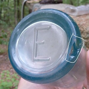Puede incluir: Una botella de vidrio transparente y antigua con un borde de color verde azulado. La parte inferior de la botella presenta una letra "E" en relieve en el centro. La botella tiene un &aacute;rea ligeramente da&ntilde;ada en el lateral.