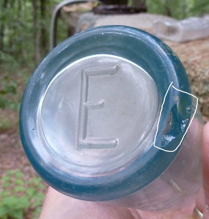Puede incluir: Una botella de vidrio transparente y antigua con un borde de color verde azulado. La parte inferior de la botella presenta una letra "E" en relieve en el centro. La botella tiene un &aacute;rea ligeramente da&ntilde;ada en el lateral.