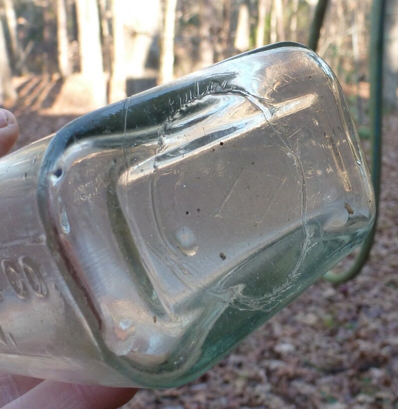 May include: Close-up of the bottom of a clear glass bottle with a green tint. The bottle has a rounded bottom and a slightly textured surface.