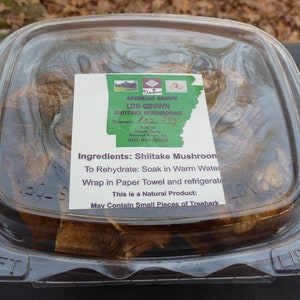 May include: A clear plastic container with a label that reads "Arkansas Grown Log Grown Shiitake Mushrooms" and "Ingredients: Shiitake Mushroom". The container holds dried mushrooms.