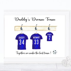 May include: A white framed print with the text "Daddy's Dream Team" and three blue soccer jerseys hanging on a wooden rack. The jerseys have the names "Daddy", "Mummy", and "Frankie" with the numbers 34, 33, and 2 on them. The text "Together we make the best team!" is below the jerseys with a soccer ball icon.