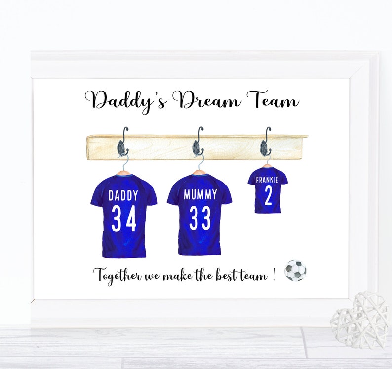 May include: A white framed print with the text "Daddy's Dream Team" and three blue soccer jerseys hanging on a wooden rack. The jerseys have the names "Daddy", "Mummy", and "Frankie" with the numbers 34, 33, and 2 on them. The text "Together we make the best team!" is below the jerseys with a soccer ball icon.