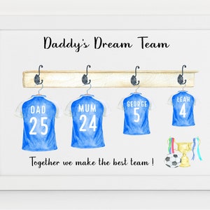 May include: A white framed print with the text "Daddy's Dream Team" and a watercolor illustration of four blue soccer jerseys hanging on a wooden rack. The jerseys have the names "Dad", "Mum", "George", and "Leah" with corresponding numbers. A soccer ball and a trophy are below the jerseys.