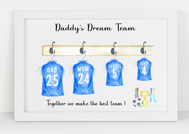 May include: A white framed print with the text "Daddy's Dream Team" and a watercolor illustration of four blue soccer jerseys hanging on a wooden rack. The jerseys have the names "Dad", "Mum", "George", and "Leah" with corresponding numbers. A soccer ball and a trophy are below the jerseys.