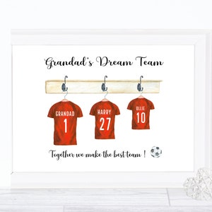 May include: A white framed print with the text "Grandad's Dream Team" and three red soccer jerseys hanging on a wooden rack. The jerseys have the names "Grandad", "Harry", and "Ollie" with the numbers 1, 27, and 10 on them. The text "Together we make the best team!" is below the jerseys with a soccer ball.