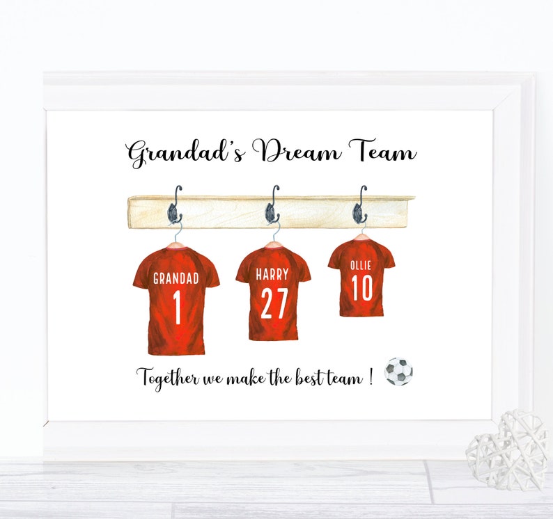 May include: A white framed print with the text "Grandad's Dream Team" and three red soccer jerseys hanging on a wooden rack. The jerseys have the names "Grandad", "Harry", and "Ollie" with the numbers 1, 27, and 10 on them. The text "Together we make the best team!" is below the jerseys with a soccer ball.