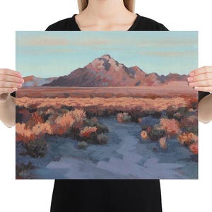 Person holding a large western landscape painting of a beautiful desert during sunset with a mountain in the background, orange peach desert bushes and plants and a trail leading to the mountain, with a light blue sky.