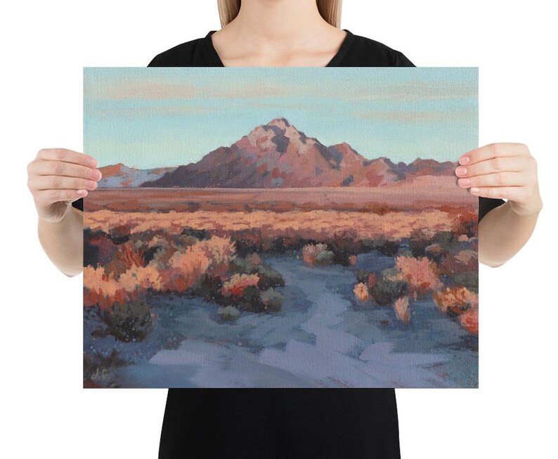 Person holding a large western landscape painting of a beautiful desert during sunset with a mountain in the background, orange peach desert bushes and plants and a trail leading to the mountain, with a light blue sky.