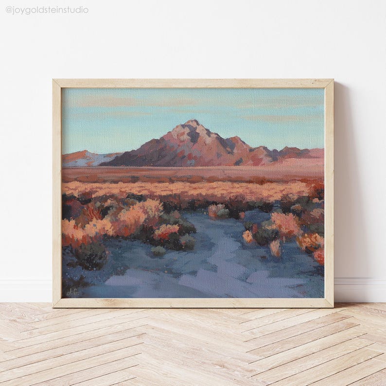 A framed southwest art print of a desert landscape with a mountain range in the background. The sky is a light blue, and the mountains are a mauve-brown color. The foreground is a field of dry, brown brush with a trail leading to the mountain.