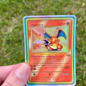 May include: A holographic Charizard Pokemon card from the first edition set. The card features a red, orange, and yellow Charizard with blue wings and a fiery background. The card has a 120 HP rating and the text "Fire Spin" and "Energy Burn".