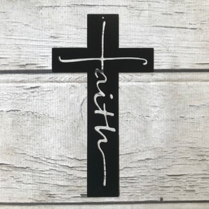 Puede incluir: Una cruz de metal negro con la palabra "faith" en fuente cursiva. La cruz est&aacute; montada sobre un fondo de madera blanca. La cruz es un s&iacute;mbolo religioso.