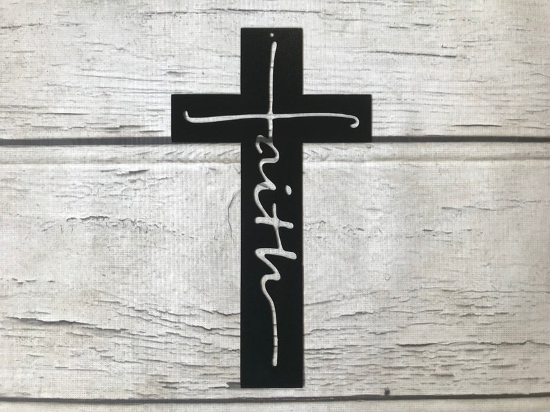 Puede incluir: Una cruz de metal negro con la palabra "faith" en fuente cursiva. La cruz est&aacute; montada sobre un fondo de madera blanca. La cruz es un s&iacute;mbolo religioso.