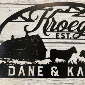 May include: Black metal sign with a farmhouse theme. It features a barn, two cows, clouds, and the text "Kroegels Est. 2017 Dane & Kane".