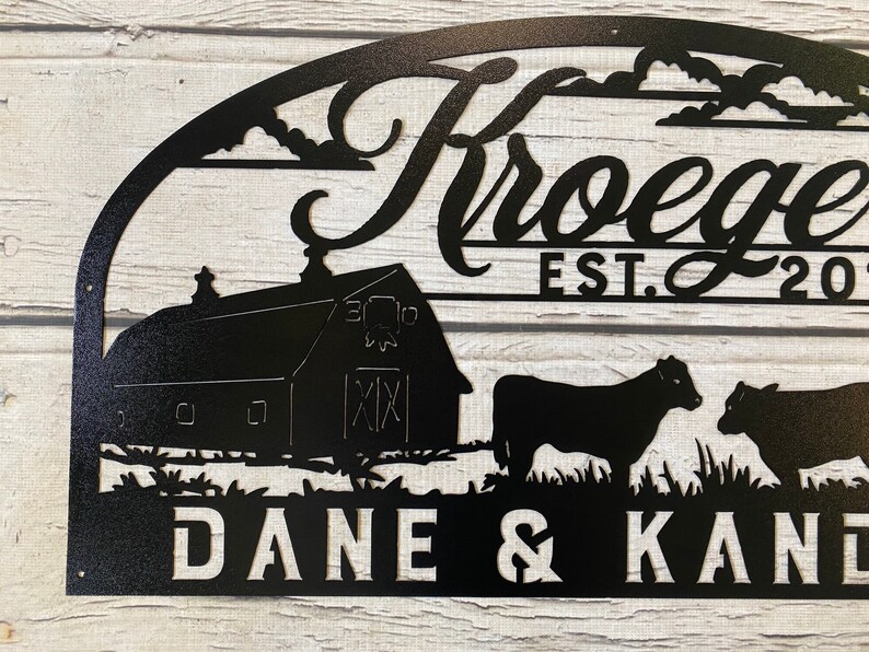 May include: Black metal sign with a farmhouse theme. It features a barn, two cows, clouds, and the text "Kroegels Est. 2017 Dane & Kane".