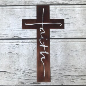 Puede incluir: Una cruz de madera marr&oacute;n con la palabra "fe" recortada en una fuente blanca y cursiva. La cruz se muestra sobre un fondo de tablones de madera blancos y grises. Un art&iacute;culo de decoraci&oacute;n religiosa.