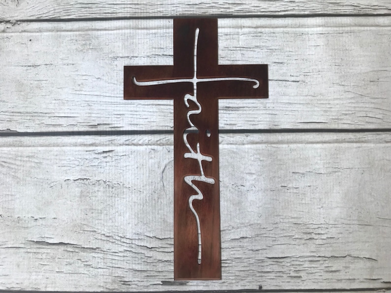 Puede incluir: Una cruz de madera marr&oacute;n con la palabra "fe" recortada en una fuente blanca y cursiva. La cruz se muestra sobre un fondo de tablones de madera blancos y grises. Un art&iacute;culo de decoraci&oacute;n religiosa.