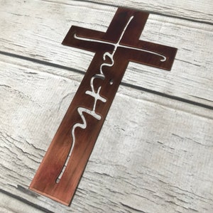 Puede incluir: Una cruz de metal r&uacute;stico con la palabra "faith" recortada en una fuente cursiva. La cruz es de color marr&oacute;n c&aacute;lido y se muestra sobre un fondo de madera clara. El dise&ntilde;o es simple y elegante, ideal para decoraci&oacute;n.