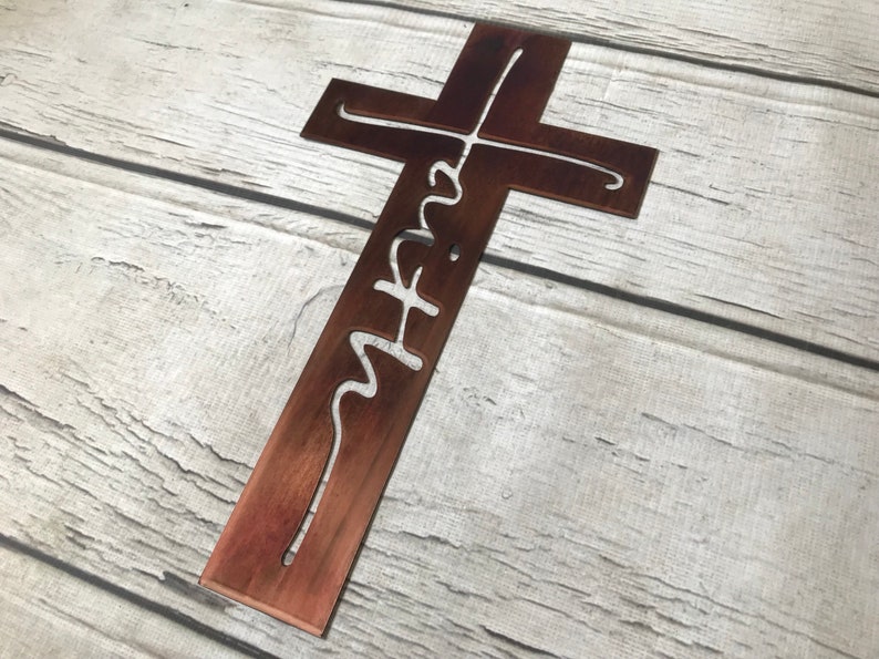 Puede incluir: Una cruz de metal r&uacute;stico con la palabra "faith" recortada en una fuente cursiva. La cruz es de color marr&oacute;n c&aacute;lido y se muestra sobre un fondo de madera clara. El dise&ntilde;o es simple y elegante, ideal para decoraci&oacute;n.