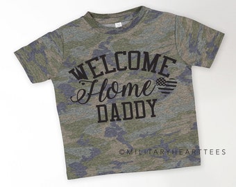 May include: Camouflage t-shirt with the text "Welcome Home Daddy" in black lettering. A small American flag is inside a heart shape.