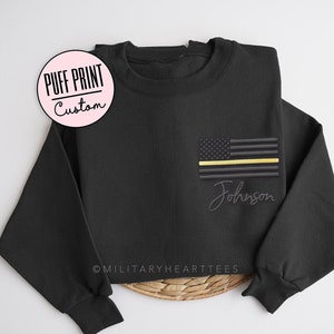 May include: Black sweatshirt with an embroidered American flag featuring a gold stripe and the name "Johnson" in script. The sweatshirt has long sleeves and a crew neck. The text "PUFF PRINT Custom" is in a pink circle.
