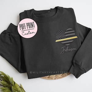 May include: Black sweatshirt with a puff print of an American flag heart with a thin yellow line and the name "Johnson" below it.