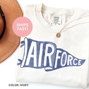 May include: Ivory t-shirt with a blue watercolor-style pennant that says "AIR FORCE".