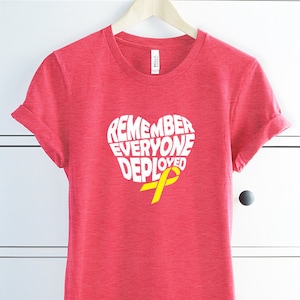 May include: Red t-shirt with a white heart-shaped graphic that says "Remember Everyone Deployed" with a yellow ribbon inside the heart. The shirt is hanging on a hanger in front of a white chest of drawers.