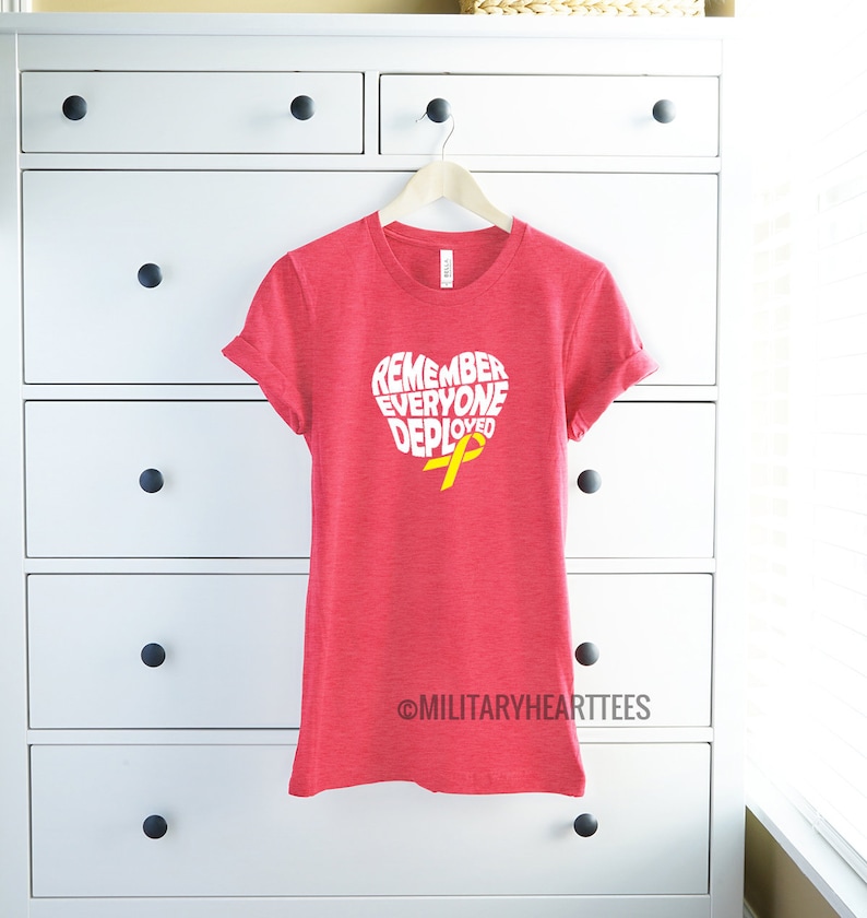 May include: Red t-shirt with a white heart-shaped graphic that says "Remember Everyone Deployed" with a yellow ribbon inside the heart. The shirt is hanging on a hanger in front of a white chest of drawers.