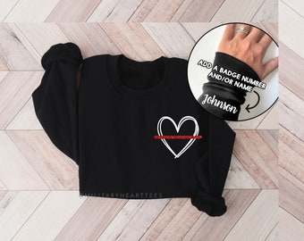 May include: Black sweatshirt with a white outline of a heart with a red line through it. The text "ADD A BADGE NUMBER AND/OR NAME" is written on a black cuff with the name "Johnson" written on it.