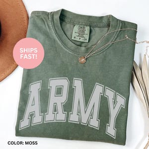 May include: A moss green t-shirt with the word "ARMY" in large, white, arched letters. The shirt has a crew neck and a small tag on the upper back. A gold necklace with a pendant rests on the shirt. The text "SHIPS FAST!" is on a pink circle.