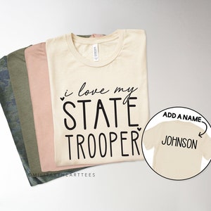 May include: A beige t-shirt with the text "i love my STATE TROOPER" and a heart symbol. A second beige t-shirt with the text "ADD A NAME" and an arrow pointing to the text "JOHNSON" is shown in a circle.