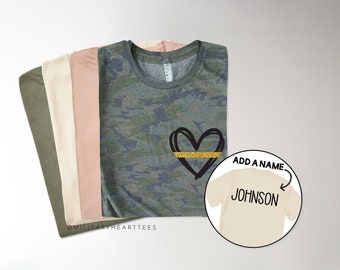 May include: A stack of t-shirts in various shades of green, beige, and pink. The top shirt features a camouflage pattern with a black heart outlined in gold glitter. The text "ADD A NAME" is written above a beige t-shirt with the name "JOHNSON" printed on it.