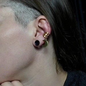 May include: A gold-colored ear cuff with a crescent moon charm and a black ear plug. The ear cuff has a layered design. The jewelry is worn on the ear.