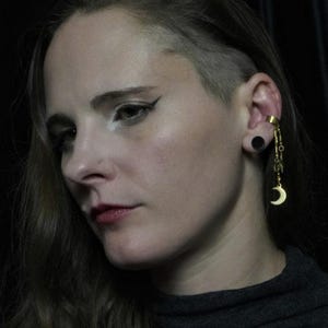 May include: A gold-colored ear cuff with a dangling chain and a crescent moon charm. The ear cuff is worn on the upper ear, and a black plug is in the earlobe. The person is wearing a gray turtleneck.