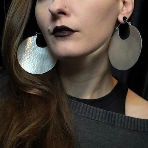 Puede incluir: Pendientes de aro grandes, de color plateado, con una textura martillada y un recorte en forma de media luna. Los pendientes son circulares y cuelgan de un alambre simple. La joya se lleva en la oreja.