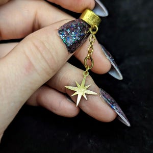 May include: A gold-colored charm with a starburst design, suspended from a small chain and a ring. The charm is attached to a gold-colored cylindrical piece. The background is dark.