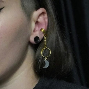 May include: A gold-colored ear cuff and chain earring with a black moon charm. The earring is attached to a gold ring and chain, and the ear has a black plug. The person has dark hair.