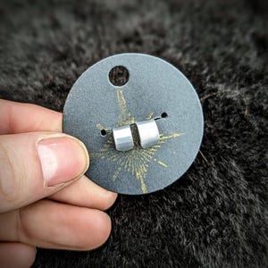 Puede incluir: Un par de pendientes de botón plateados y geométricos, exhibidos en una tarjeta circular gris oscuro. Los pendientes son pequeños cilindros rectangulares. La tarjeta tiene un agujero en la parte superior y un diseño de estrella dorada.